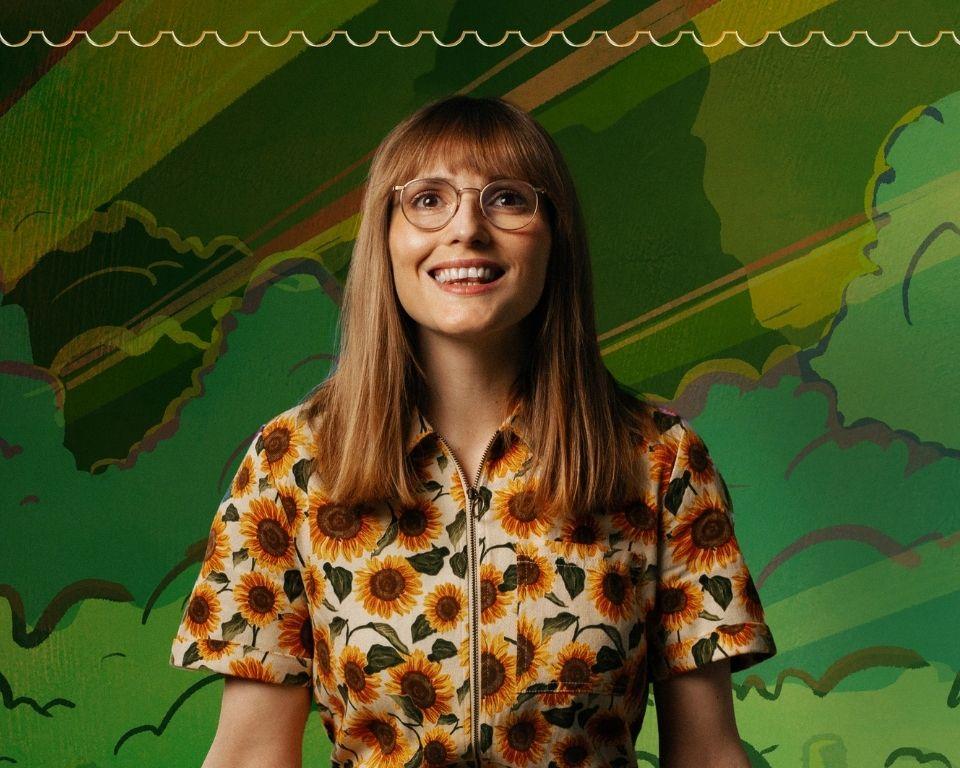 A smiling woman with long light brown hair and glasses sits against a vibrant, illustrated green background of stylised trees, clouds, and a bus. She wears a sunflower-print zip-up dress and appears cheerful and relaxed in the centre of the frame.