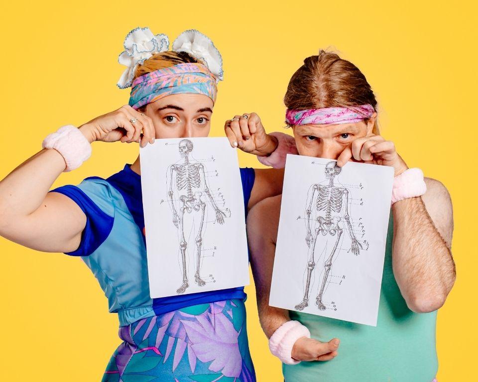 Colourful promotional image of two performers in retro fitness costumes holding anatomical skeleton diagrams. Quirky, inclusive theatre show about disability, bodies, and movement. Bold yellow background, 80s-inspired fashion, and comedy performance imagery.