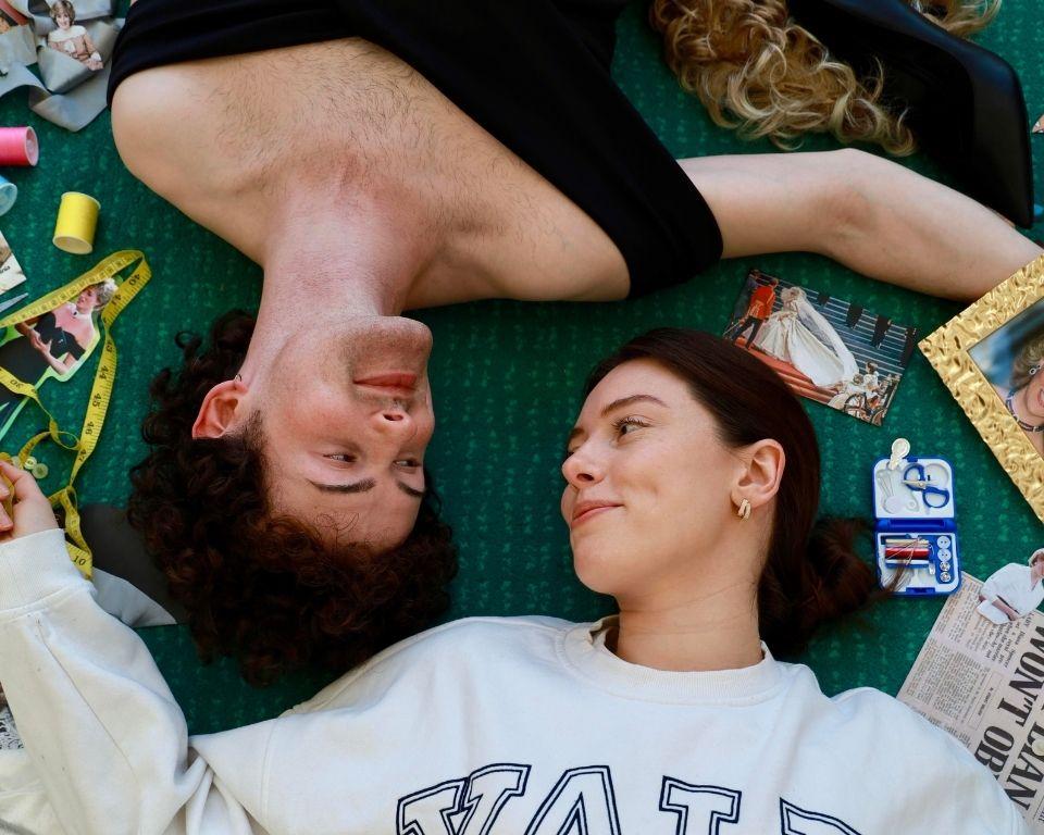 A man and woman relax on the floor surrounded by random objects, looking at each other's eyes.