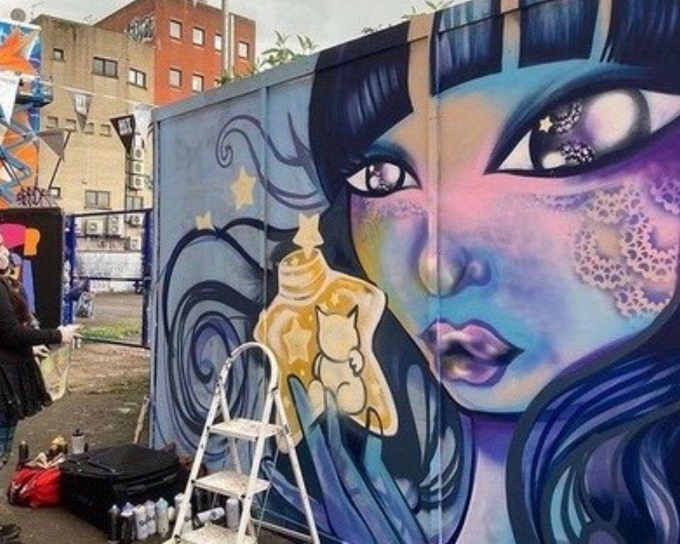 A colorful mural featuring a woman with blue hair holding a yellow bottle, surrounded by vibrant street art elements