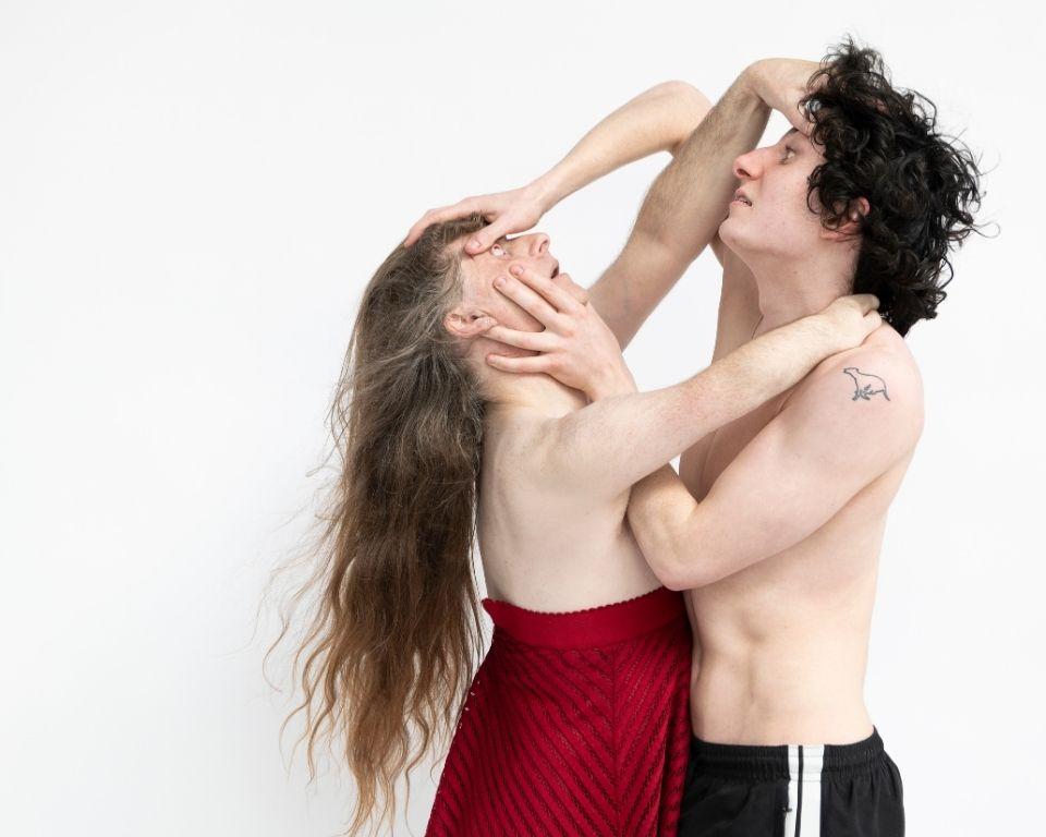 Two individuals in a dramatic, physical embrace against a white background; one with long hair in a red skirt looks upward as the other, shirtless in black shorts, holds their head and face in an intense, performance-like gesture.