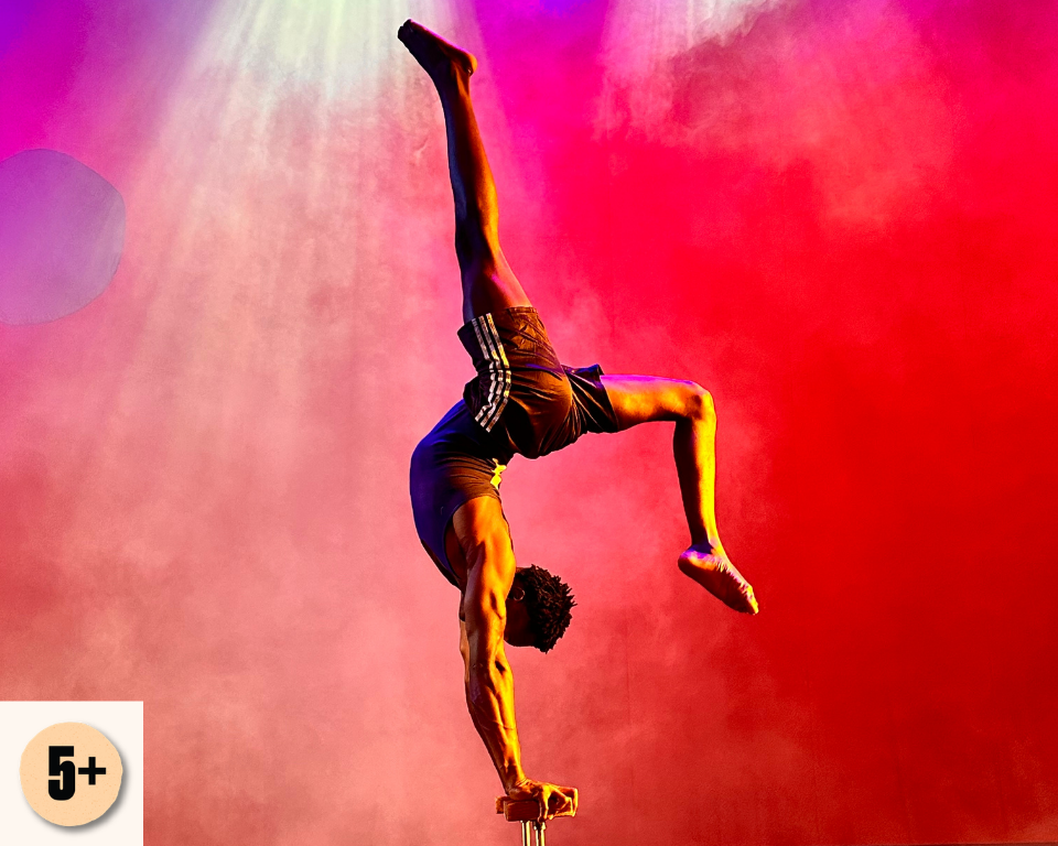 A circus performer balances in a one-handed handstand against a red and pink smoke-filled stage background. Visible access symbol: 5+.