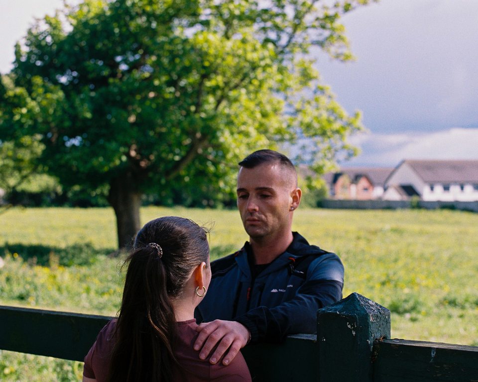 Two people standing by a wooden fence outdoors, with one person placing a hand on the other's shoulder against a backdrop of a grassy field and a large tree.