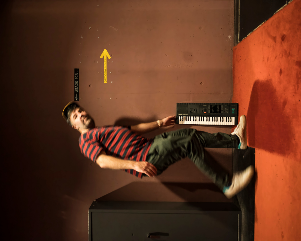 A sideways-oriented photograph of a man in a striped shirt appearing to float horizontally next to a wall keyboard backstage.