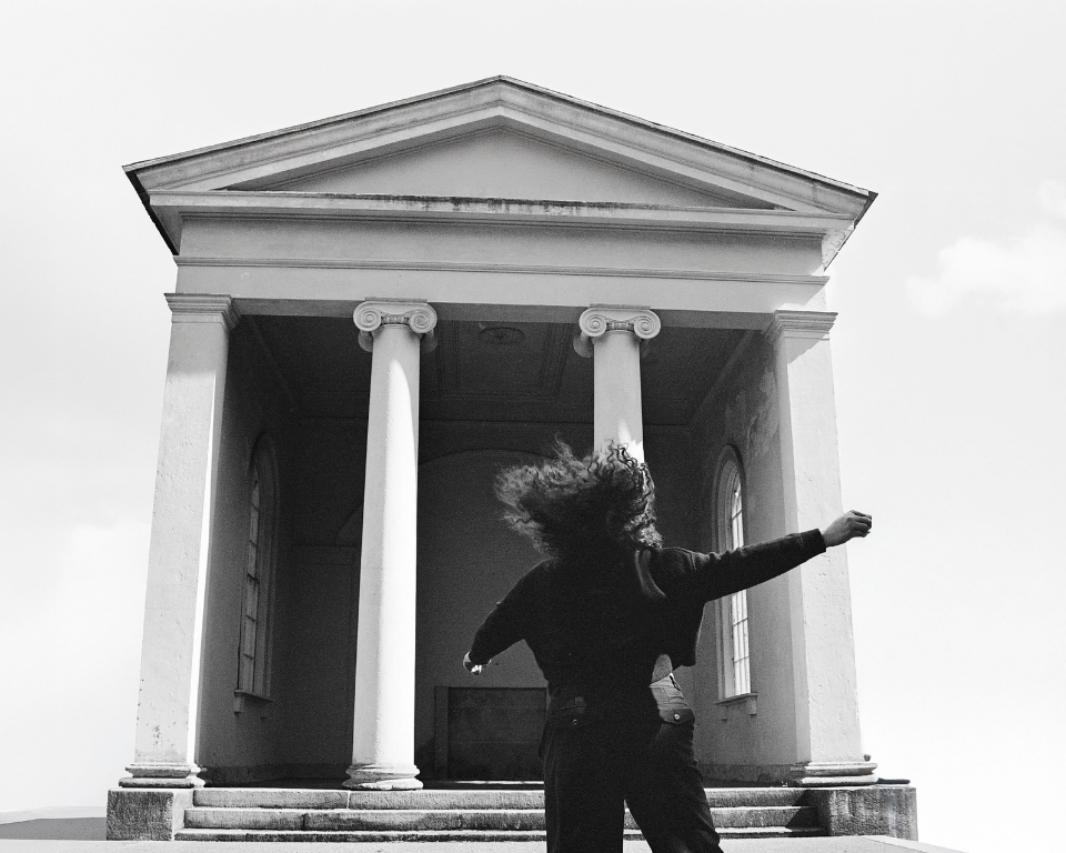 A person with arms outstretched stands before a neoclassical building with tall white columns in a dramatic black-and-white photograph.