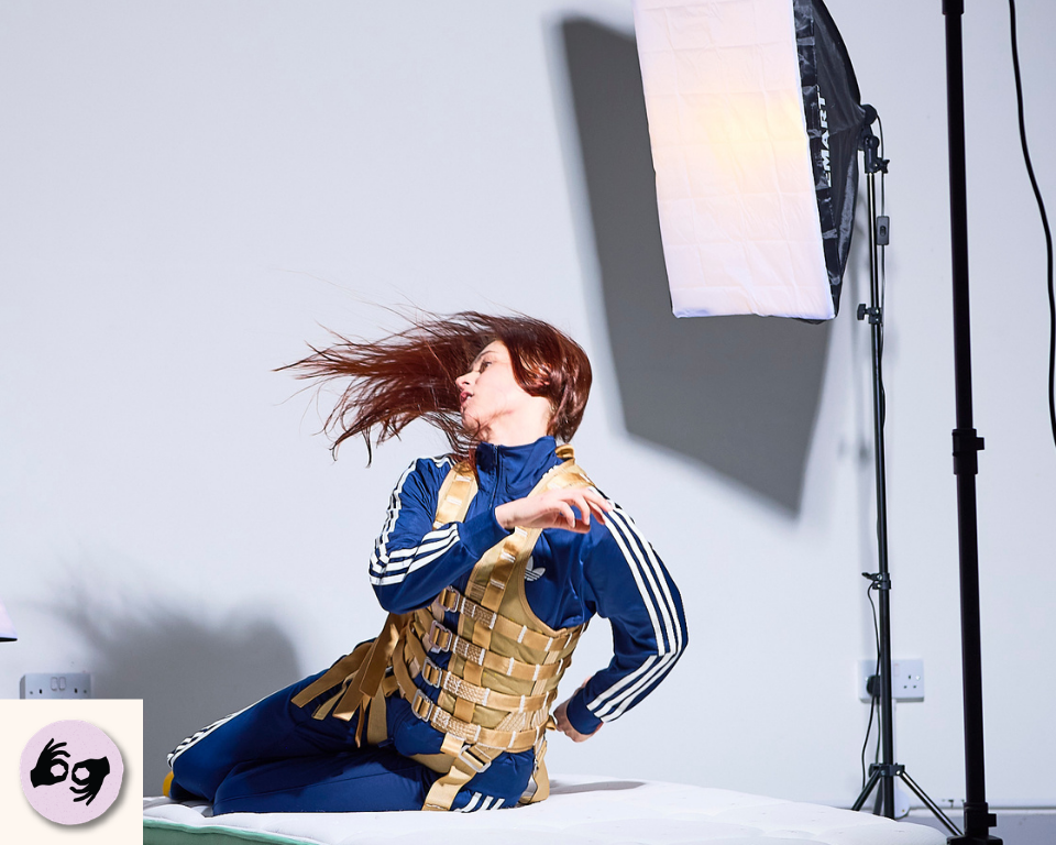 A performer in a blue tracksuit kneels on a white studio floor, turning their head as their hair moves. Visible logo: ISL.