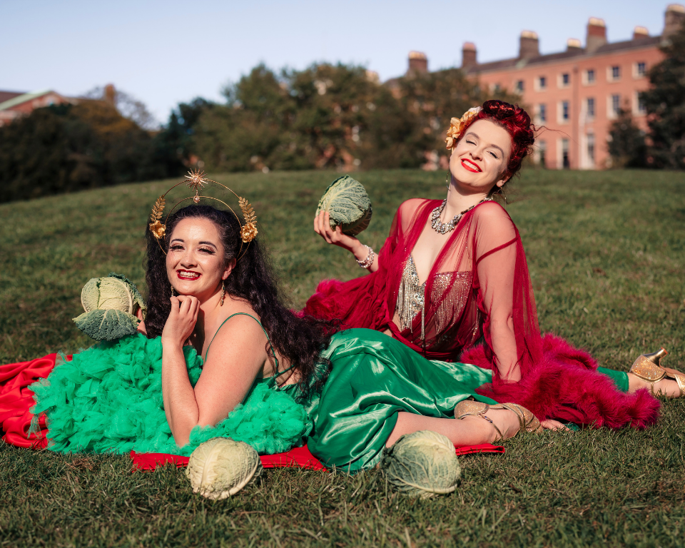 Two performers in bright red and green cabaret-style outfits pose on grass holding cabbages, with a large building in the background.