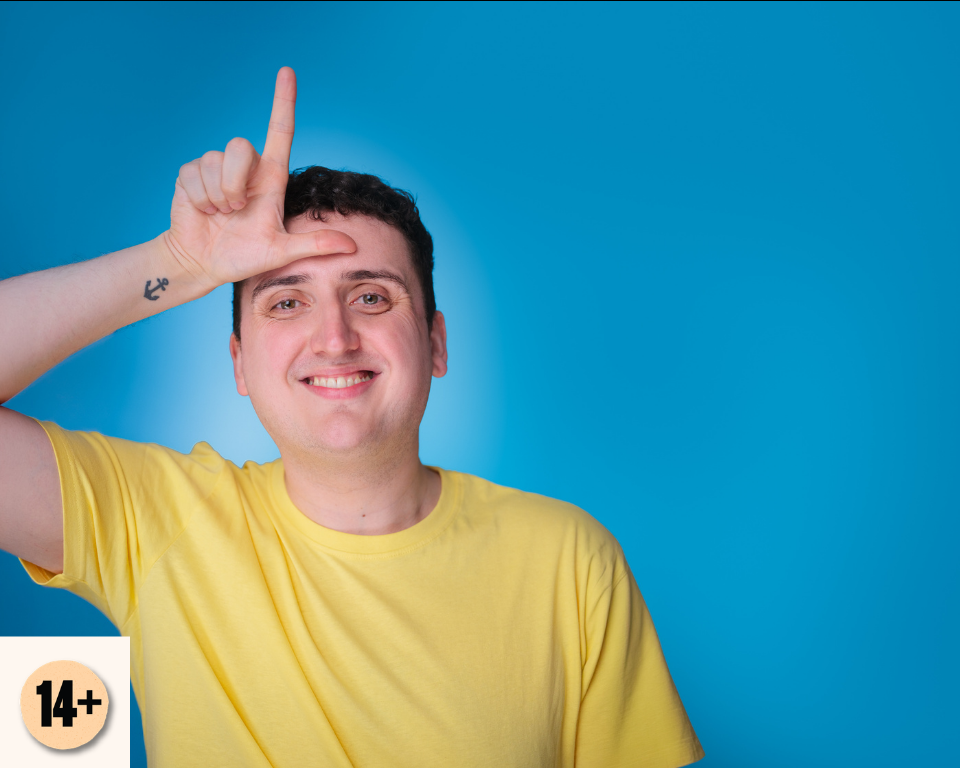 A smiling man in a yellow T-shirt makes an L shape with his hand against a bright blue studio background. Visible access symbol: 14+.