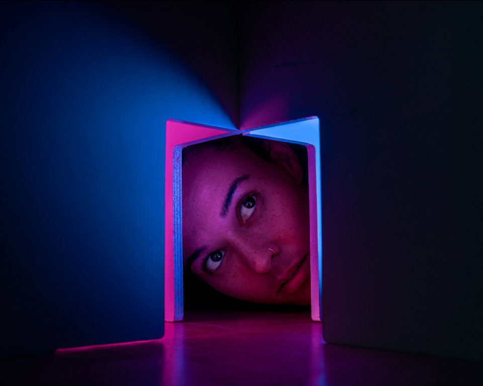 A close-up of a person’s face partly framed inside a small box, lit with pink and blue light in a dark space.