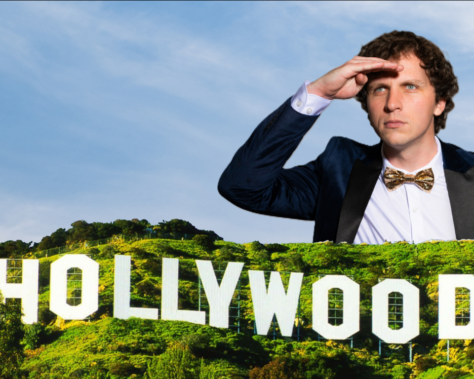 A man in a tuxedo and gold bow tie salutes while standing behind large white Hollywood-style letters on green hills.