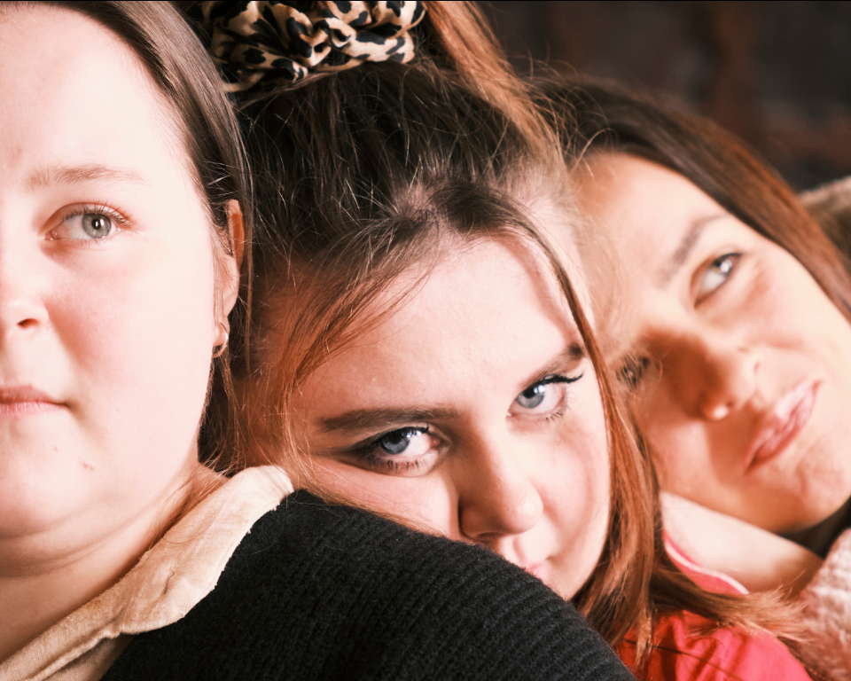 A close-up of three people leaning in tightly together, looking toward the camera with expressive facial expressions.