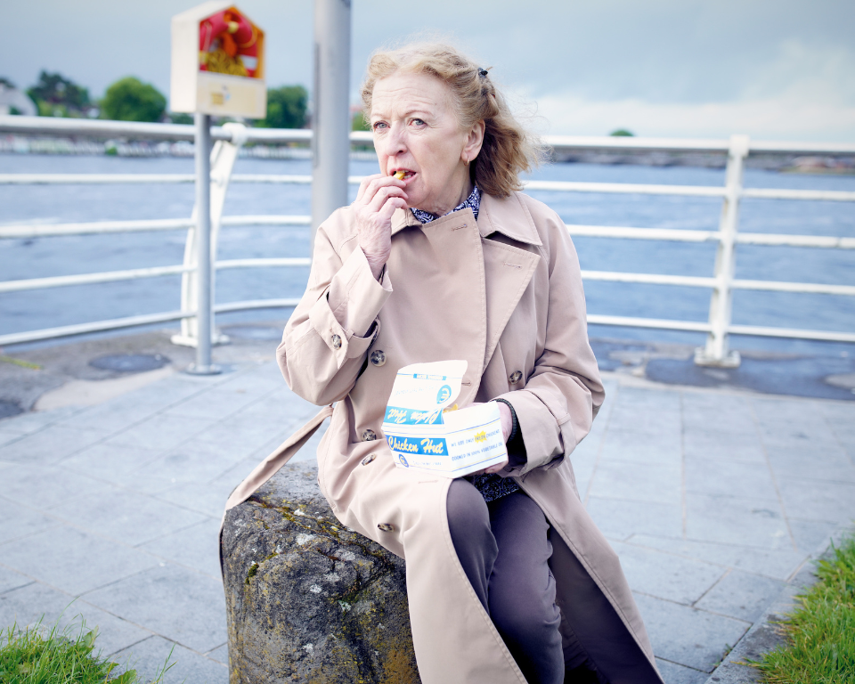 An older person wearing a beige trench coat sits outdoors near water, eating food from a takeout box.