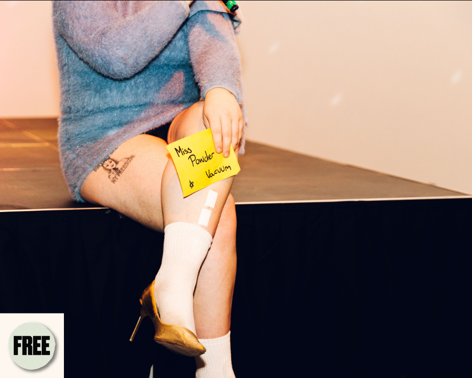 A seated performer in a blue dress and gold heel holds a yellow note reading ‘Miss Powder Vacuum’ on their leg. Visible symbol: Free.