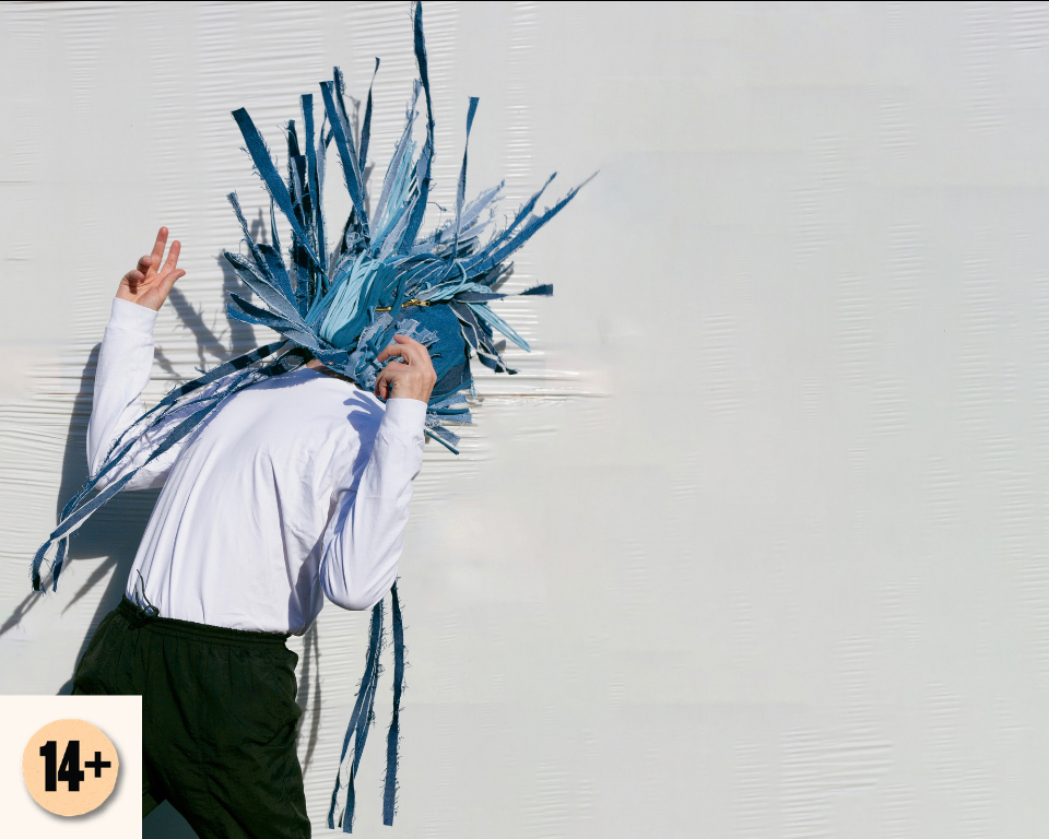 A performer with a blue shredded headpiece bends forward against a pale textured wall. Visible symbol: 14+.