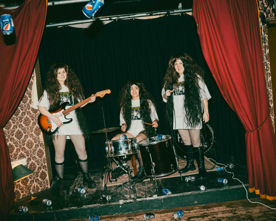 Three performers with long wigs pose as a band on a small stage with red curtains, instruments and crushed cans scattered around them.