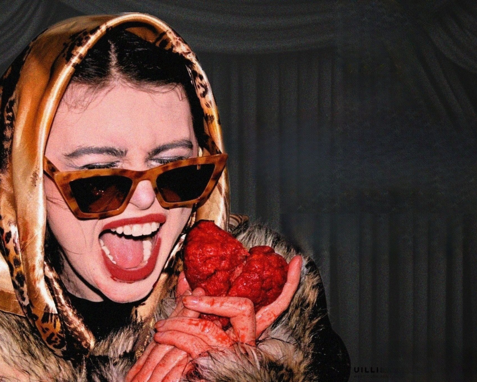 A close-up of a smiling performer in sunglasses and a patterned headscarf holding a red heart-shaped object.