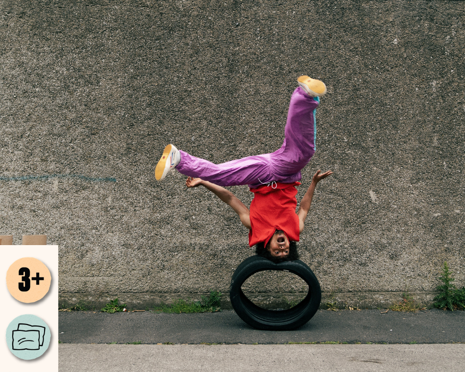 A performer in a red top and purple trousers balances upside down on a tyre against a concrete wall, with one foot kicked high in the air. Access and audience icons shown: 3+ and Relaxed Performance.