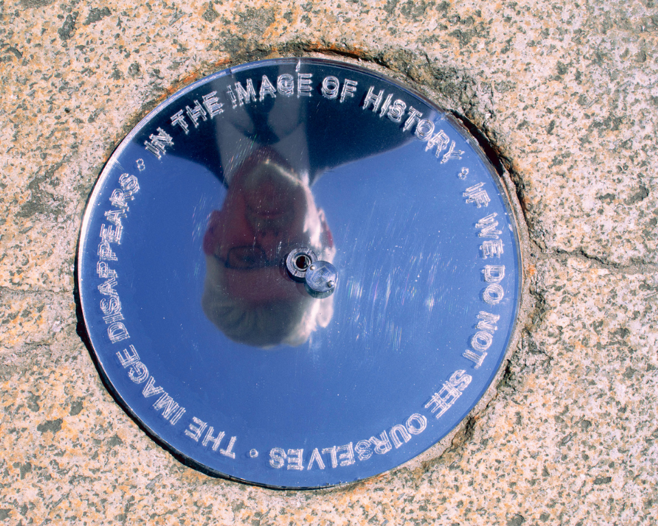 A reflective metal disc set into a stone surface shows an upside-down reflection of a person. Text around the disc reads: “The image disappears in the image of history if we do not see ourselves.” No access or audience icons shown.
