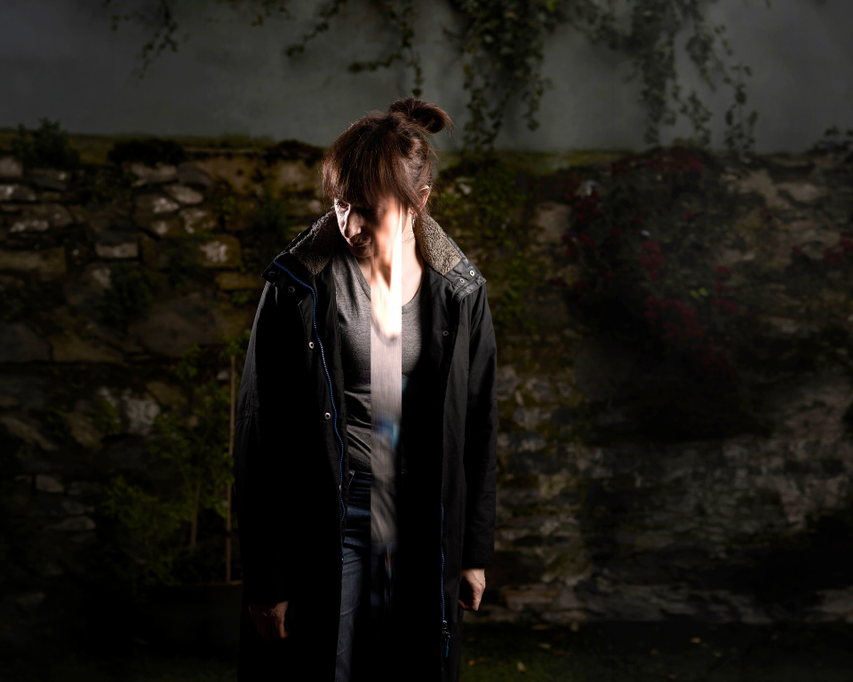 A woman stands in a dark outdoor space with a beam of light passing down the centre of her face and body.