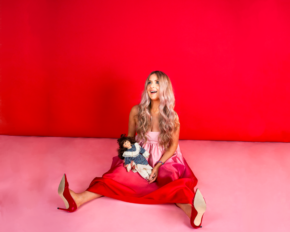 A woman in a pink dress sits against a red and pink studio background, holding a doll in her lap.