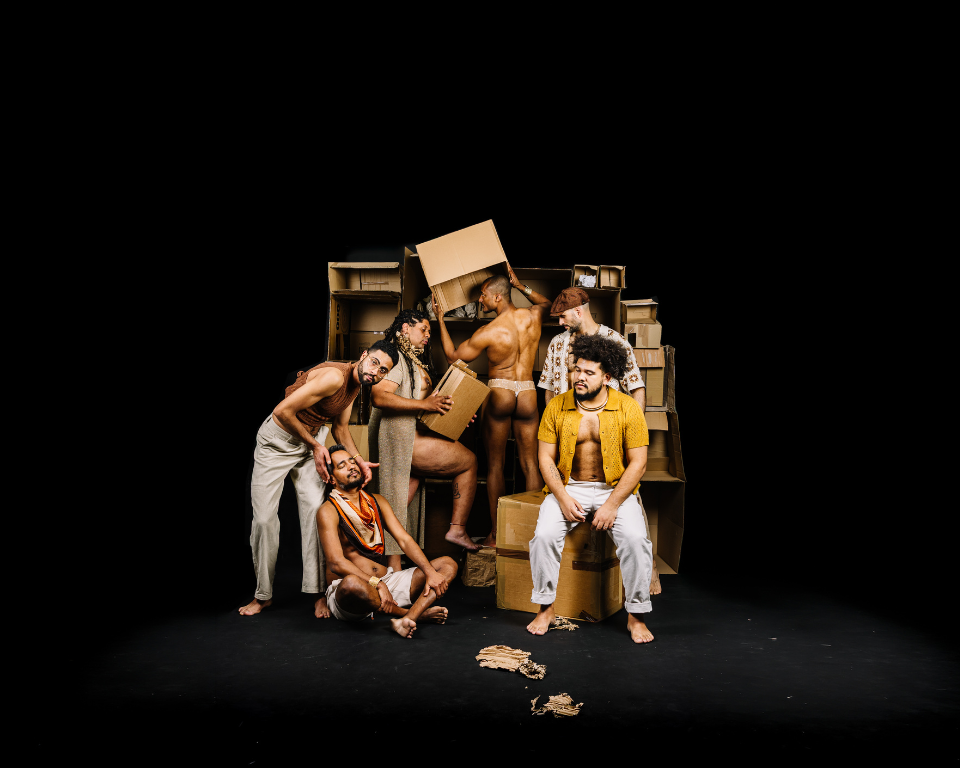 A group of performers gather around stacked cardboard boxes on a dark stage.