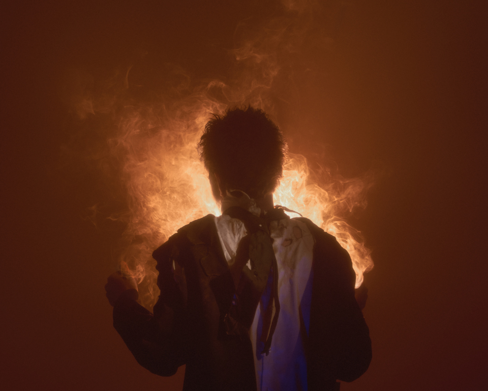 A silhouetted performer stands with bright orange smoke or fire-like light glowing behind them.