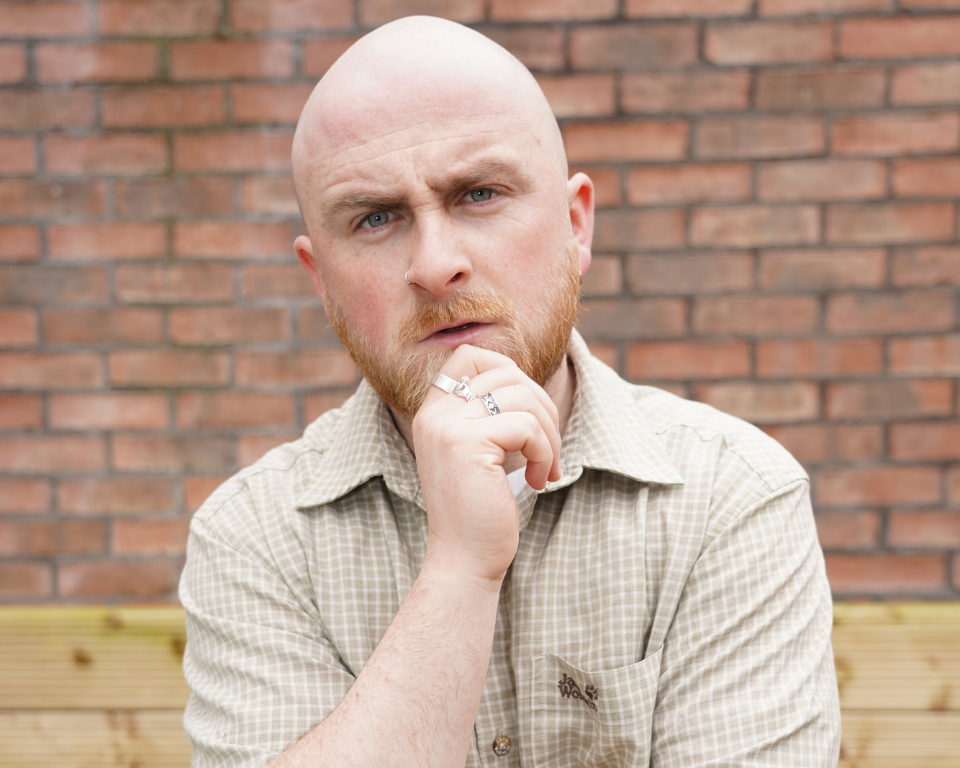 A close-up portrait of a bald, bearded man with his hand on his chin in a thinking pose against a brick wall.