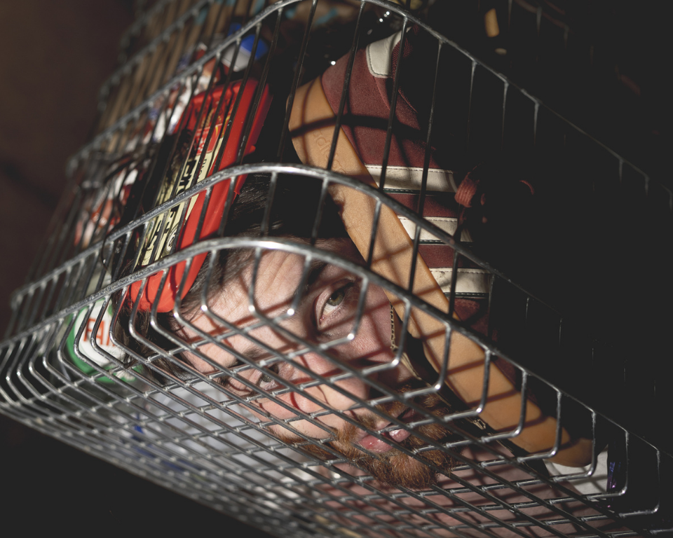 A close-up of a person’s face partly hidden behind the metal bars and shadows of a shopping trolley.