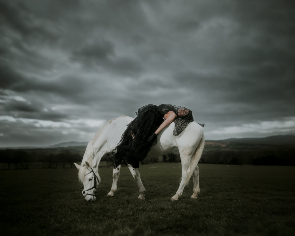 A woman draped over a white horse in a dark rural landscape under heavy grey clouds.