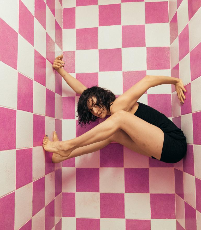 A woman in a black unitard holds herself suspended between pink and white tiled walls