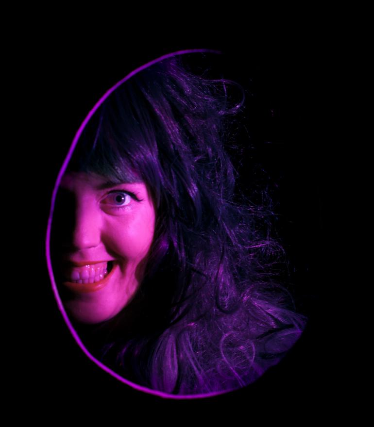A woman's smiling face, lit in purple, is visible through a oval hole in a black space