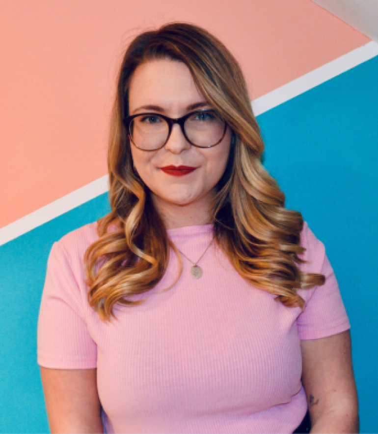 A woman wearing a pink top and glasses stands against a blue and orange background