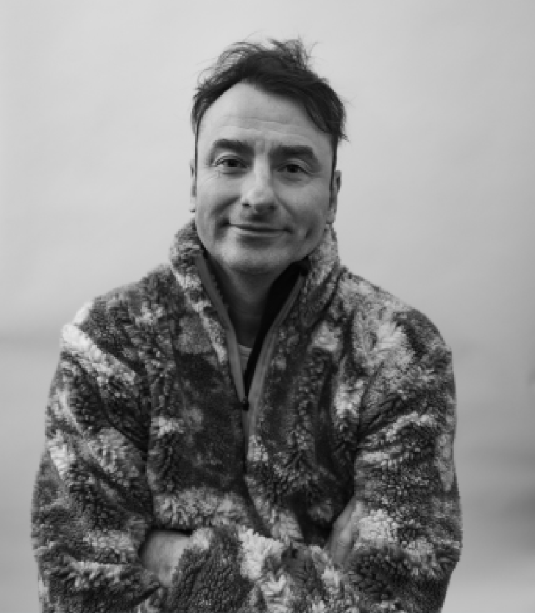 Man in a fuzzy, patterned jacket smiles subtly with arms crossed against a plain background. The monochrome tone adds a warm, relaxed feel.