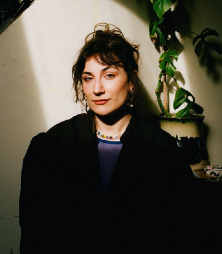 Portrait of Alba Molina Cañamero standing indoors in dramatic sunlight, wearing dark clothing, with a trailing green plant beside her.