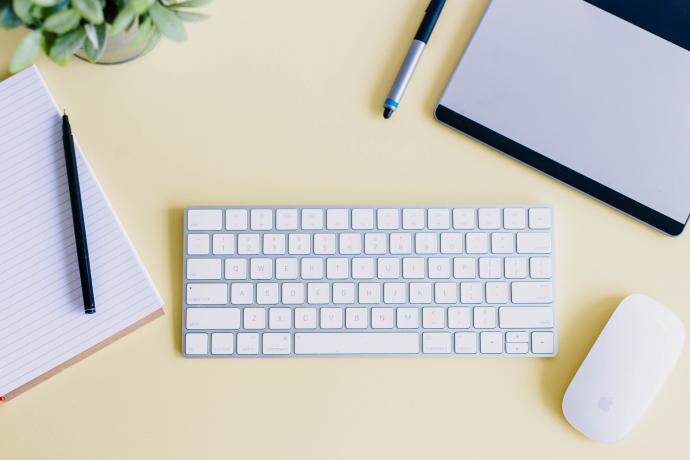 A keyboard, mouse, notebook, laptop and houseplant on a desk.