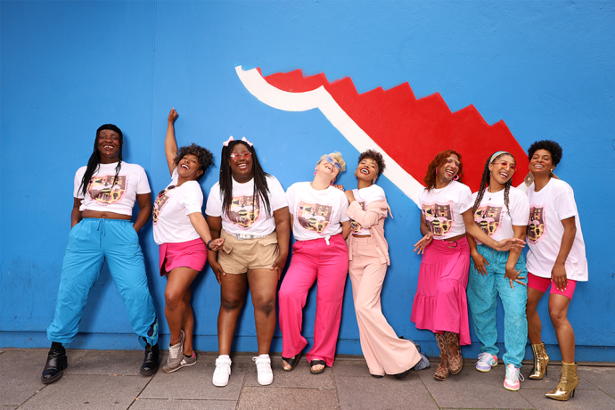 8 women of colour pose in front of a blue wall with half a painted mural
