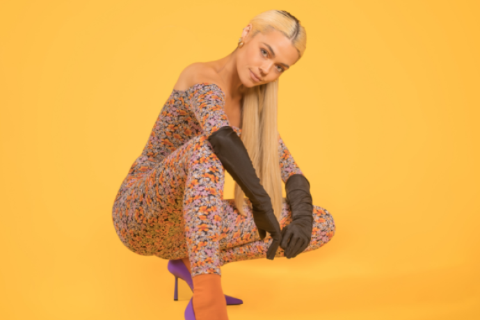 A blond woman in a body suit, high heels and elbow length gloves crouches in front of a yellow background