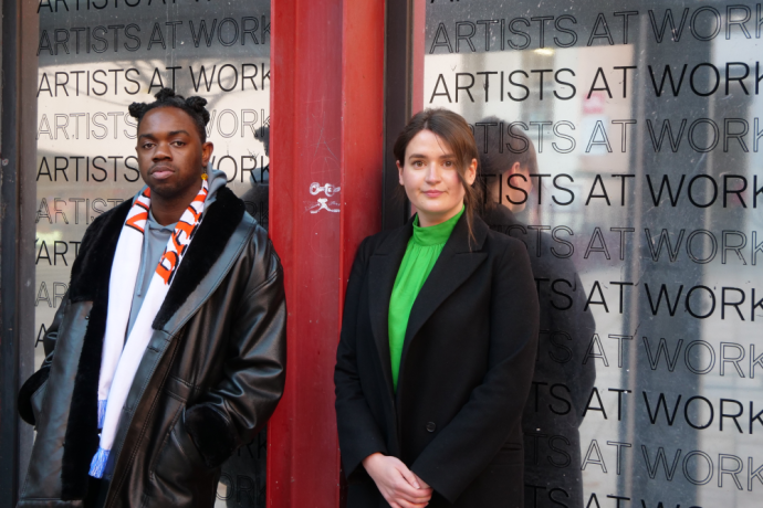 Dafe Orugbo (left) and Shanna May Breen (right) look to camera in front of a silver vinyl that say Artists At Work