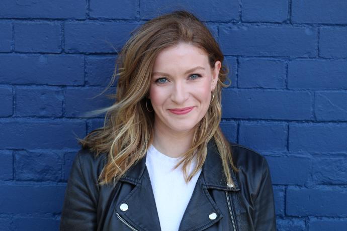 White woman with long red hear wearing a black leather biker jacket stands against a deep blue wall