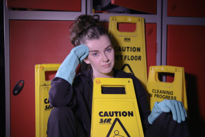 A young woman wearing blue cleaning gloves surrounded by 'slippery when wet' warning signs.