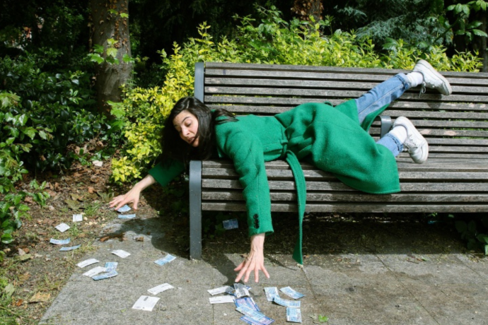 A woman lies on a park bench grabbing at scratch cards scattered on the ground. 