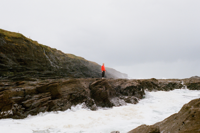 A person wearing a bright red top standing on a cliff with waves foaming below. 