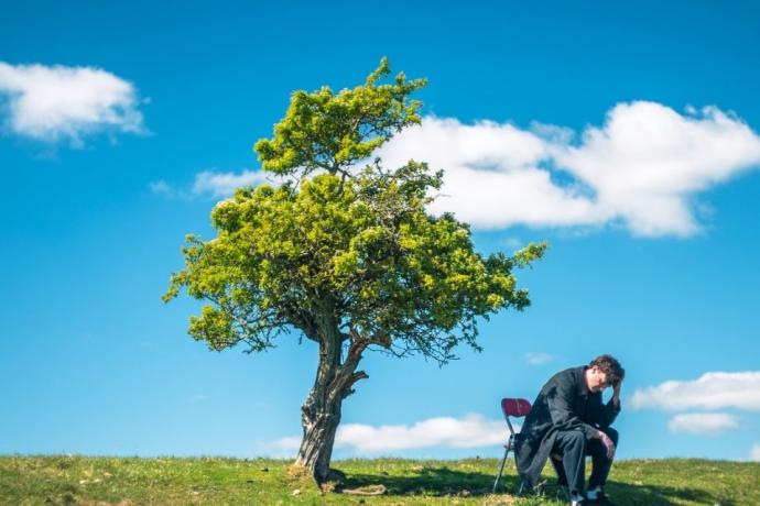A man sits on a grassy hill beside a tree, enjoying the view of the landscape around him.