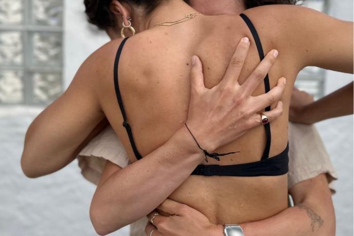Two women embrace each other while wearing matching black bikinis, smiling joyfully in a sunny outdoor setting.