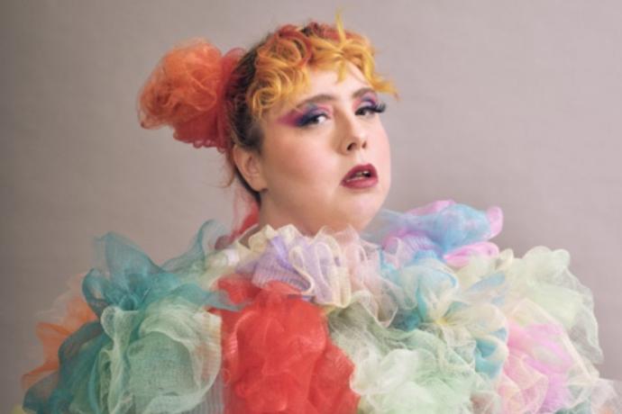 A woman with vibrant, multicolored hair wearing a bright, patterned dress stands confidently, showcasing her unique style.
