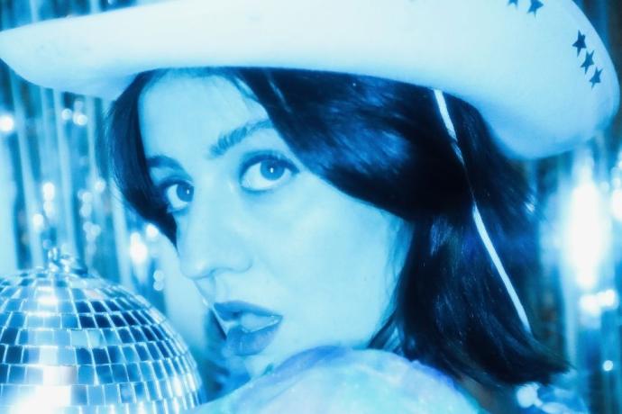 A woman wearing a cowboy hat stands beside a shimmering disco ball, blending country and disco themes.