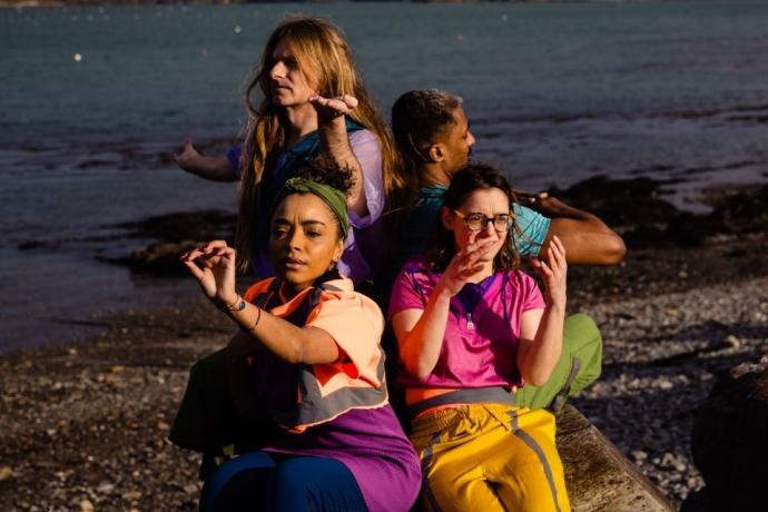 Three women in vibrant outfits sit on rocks by the water, enjoying a sunny day and the scenic view around them.