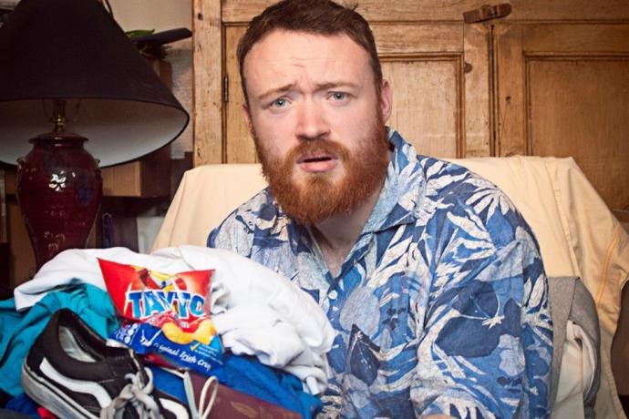 A man with a beard wearing a blue shirt sits in a chair next to a large pile of clothes.