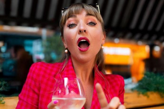 A woman in a red jacket raises a glass of white wine, outside a bar.