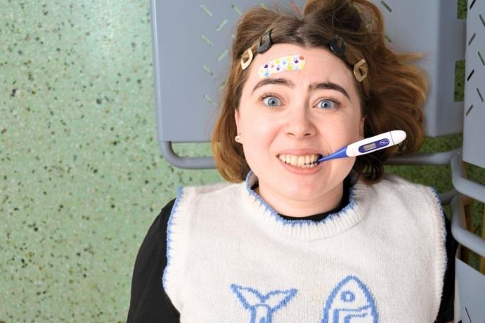 A person lies down in a medical setting, wearing a light sweater with fish designs, while a thermometer is positioned near their mouth.